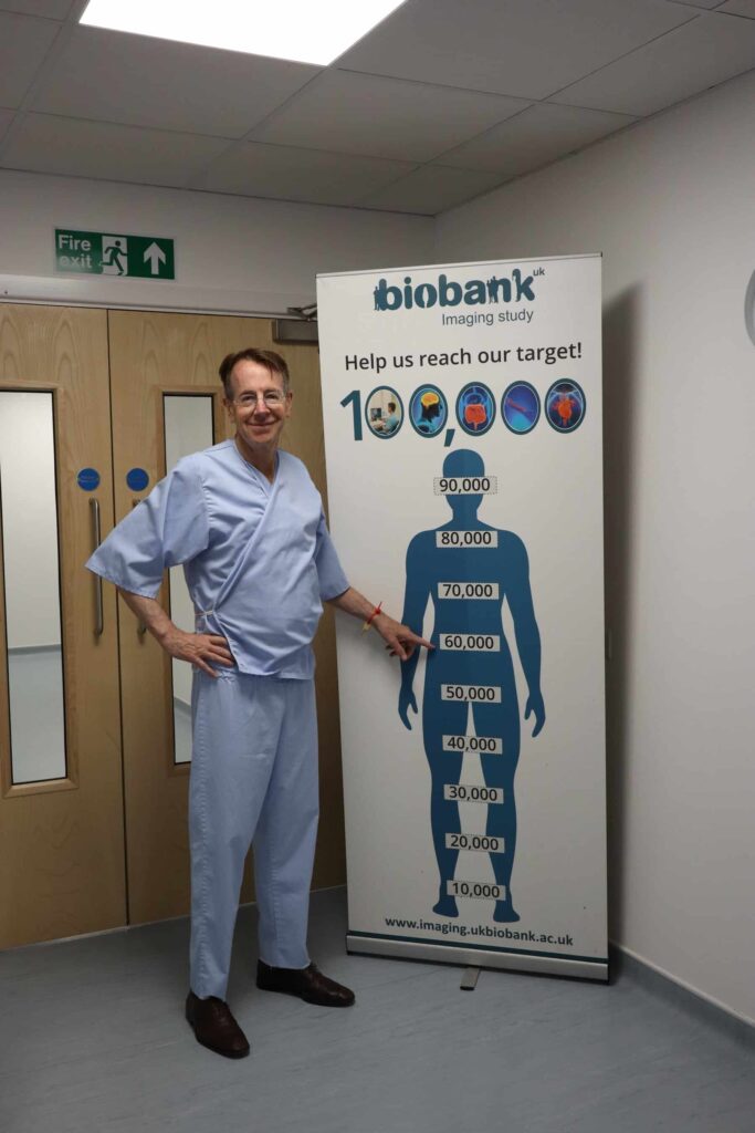 Professor Sir Rory Collins pointing to 60,000 on a banner marking the target of 100,000 imaging participants