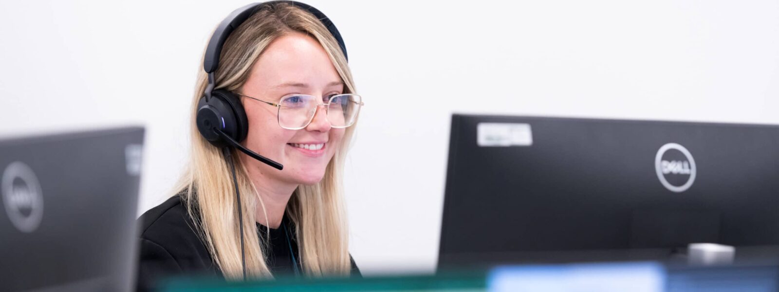 A woman sits at a desk smiling at her computer, wearing a headset