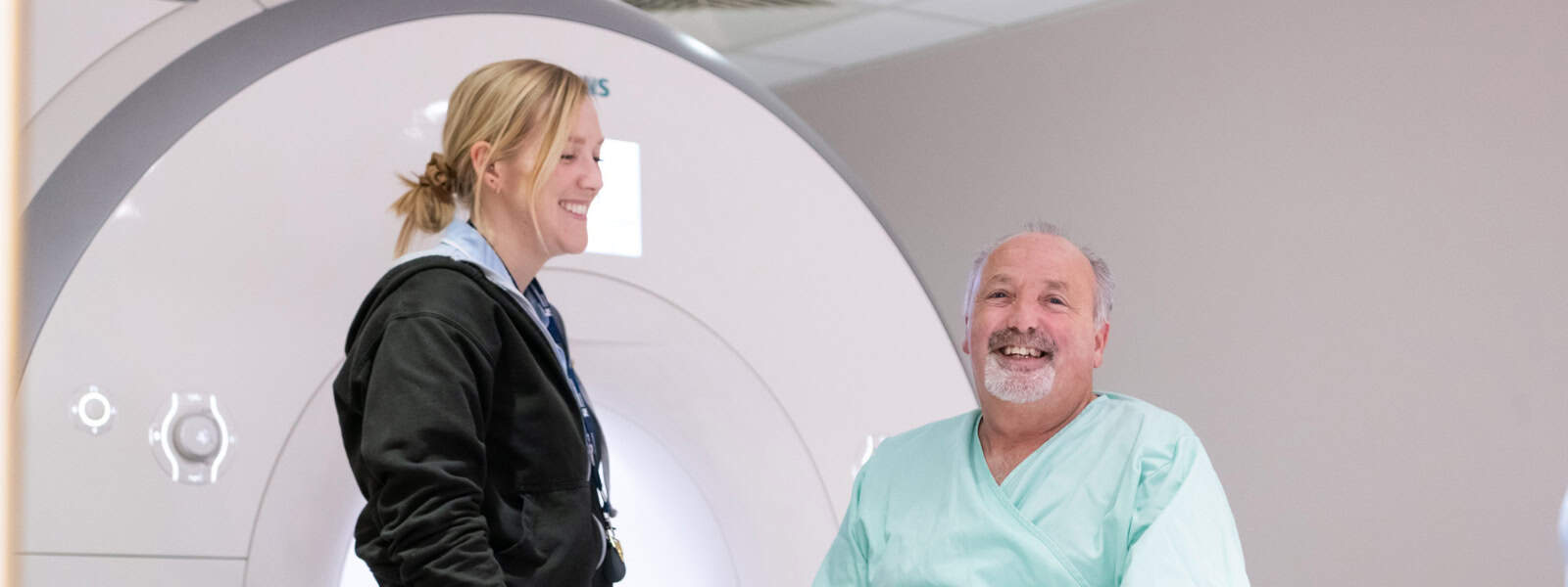A man smiles to camera in front of an MRI scanner whilst a staff member smiles at him