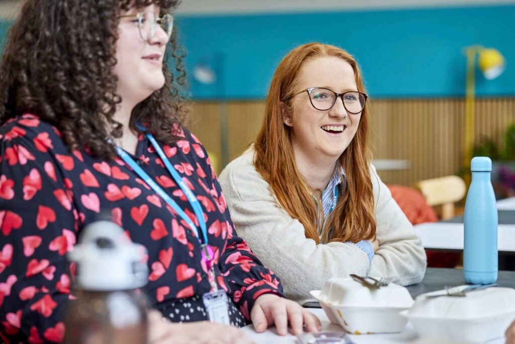 A woman sits at a table and smiles towards another colleague